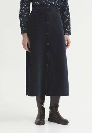 Personne portant une jupe midi noire boutonnée, une chemise à manches longues à motifs et des bottes noires montantes, debout sur un fond blanc.