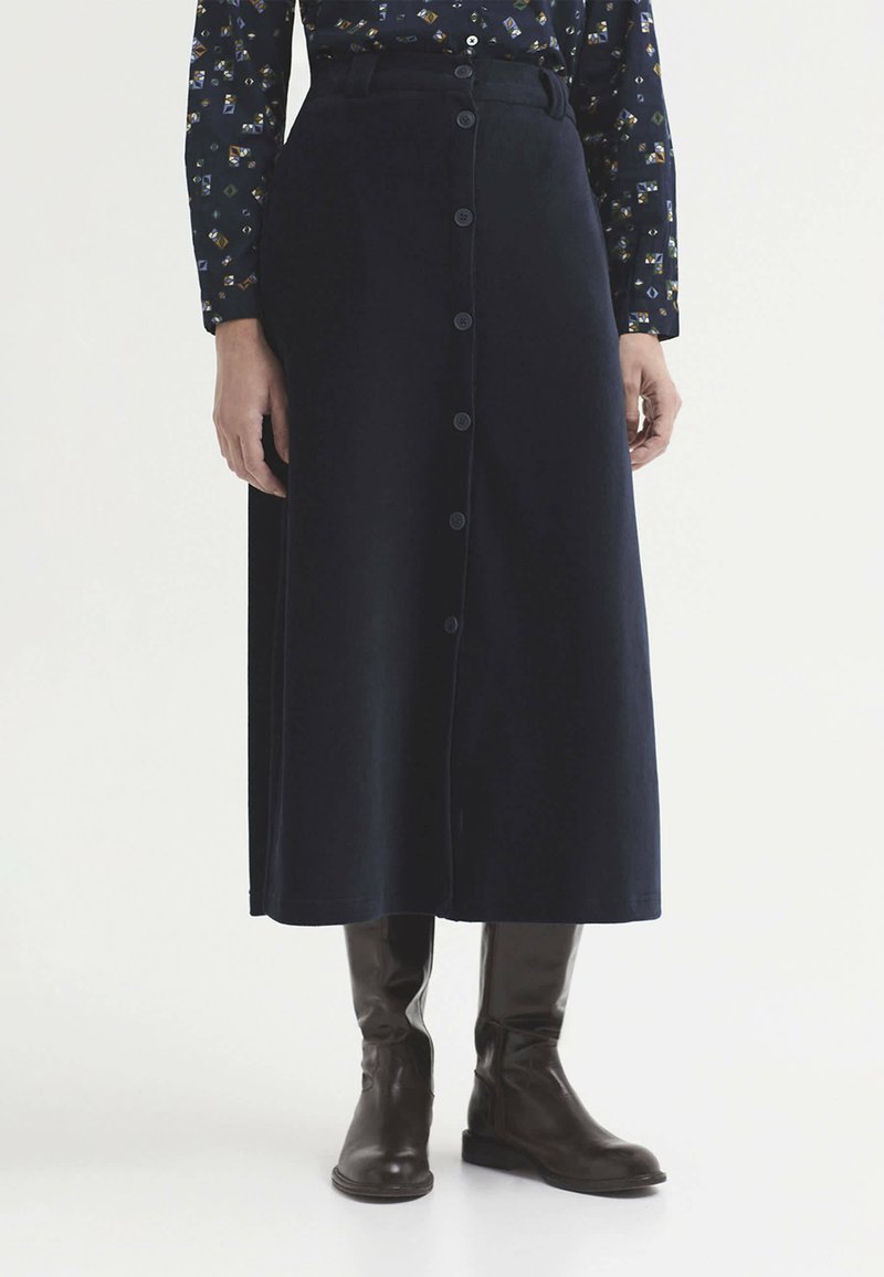 Personne portant une jupe midi noire boutonnée, une chemise à manches longues à motifs et des bottes noires montantes, debout sur un fond blanc.