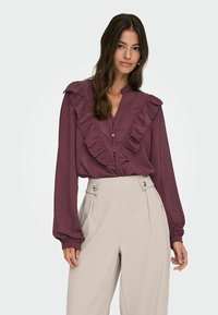 Blouse bourgogne avec devant à volants, texture à pois et fermeture à boutons, associée à un pantalon crème taille haute avec des boutons décoratifs.