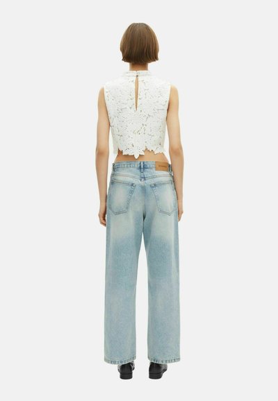 Personne debout, dos à la caméra, portant un crop top en dentelle blanche florale sans manches, un pantalon large en jean bleu clair et des chaussures noires.
