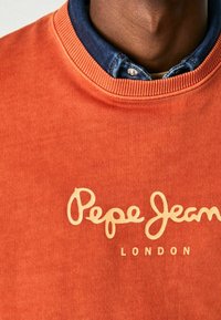 Oranžová mikina z měkké tkaniny. Má ribovanou límec, béžový kurzivní nápis "Pepe Jeans" a text "LONDON" na přední straně.