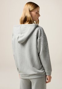 Sudadera con capucha gris, confeccionada en material suave, con adornos de rhinestones en los hombros y la parte superior de la espalda. Diseño de corte relajado.