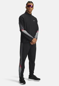 Black athletic jacket with gray and red accents, paired with matching pants. Notable striped pattern on sleeves and sides. Thick sole sneakers.