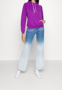 Lila huvtröja med en framficka och vita dragsnören, i kombination med ljusblå utsvängda jeans. Modellen bär vita sneakers.