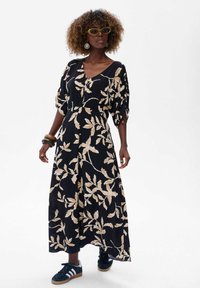 Zwarte maxi jurk met een beige bladpatroon, korte bolle mouwen en een geraffineerde taille. Draagt met zwarte sneakers en grote hoepel oorbellen.