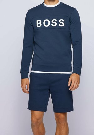 Mann trägt dunkelblauen Pullover mit dem Schriftzug "BOSS" und passende dunkelblaue Shorts vor einem einfarbigen hellen Hintergrund.