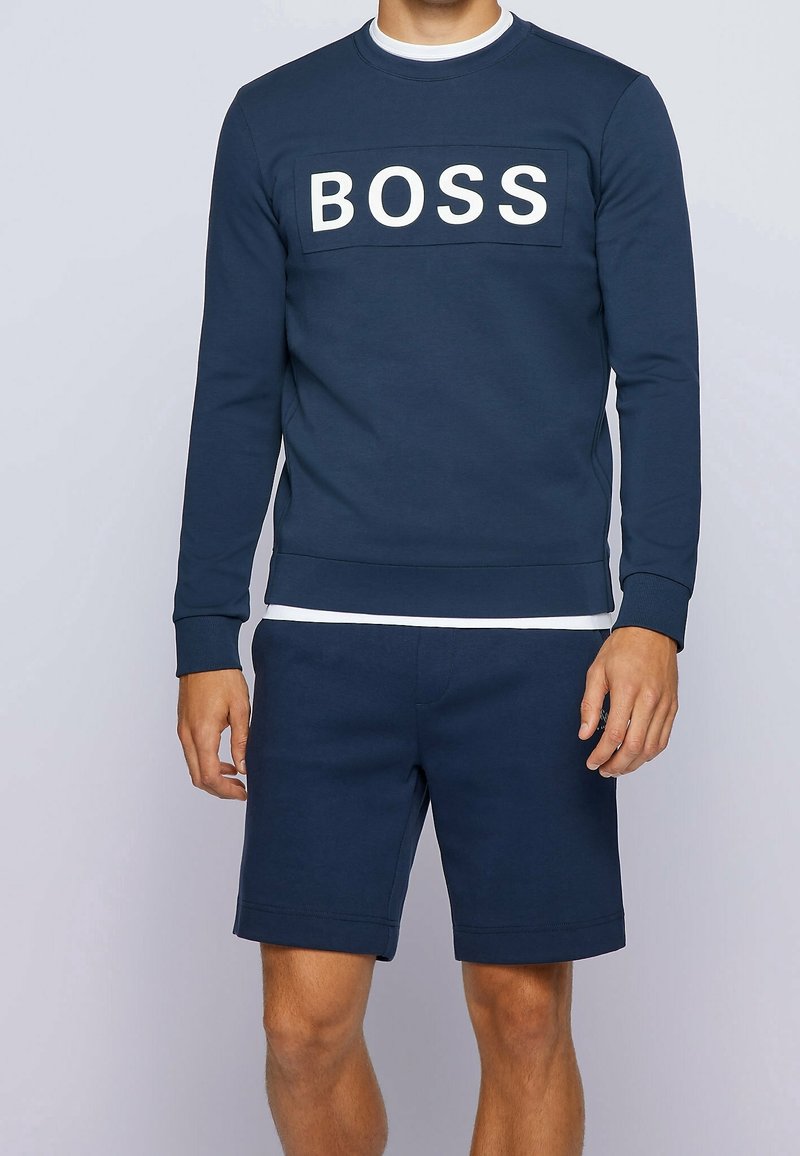 Homme portant un sweat-shirt bleu marine avec le texte "BOSS" et un short bleu marine assorti sur un fond clair uni.