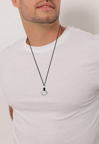 Sølv cirkulært vedhæng på en sort snor, med en tekstureret overflade og en lille knude detalje. Båret over en hvid t-shirt.