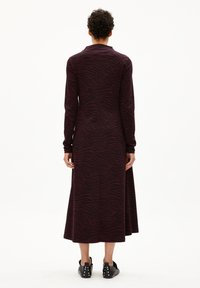 Eine Person steht mit dem Rücken zur Kamera und trägt ein langärmeliges, knöchellanges dunkles Kleid mit einem weinroten Zebramuster und schwarzen, mit Nieten besetzten Schuhen.