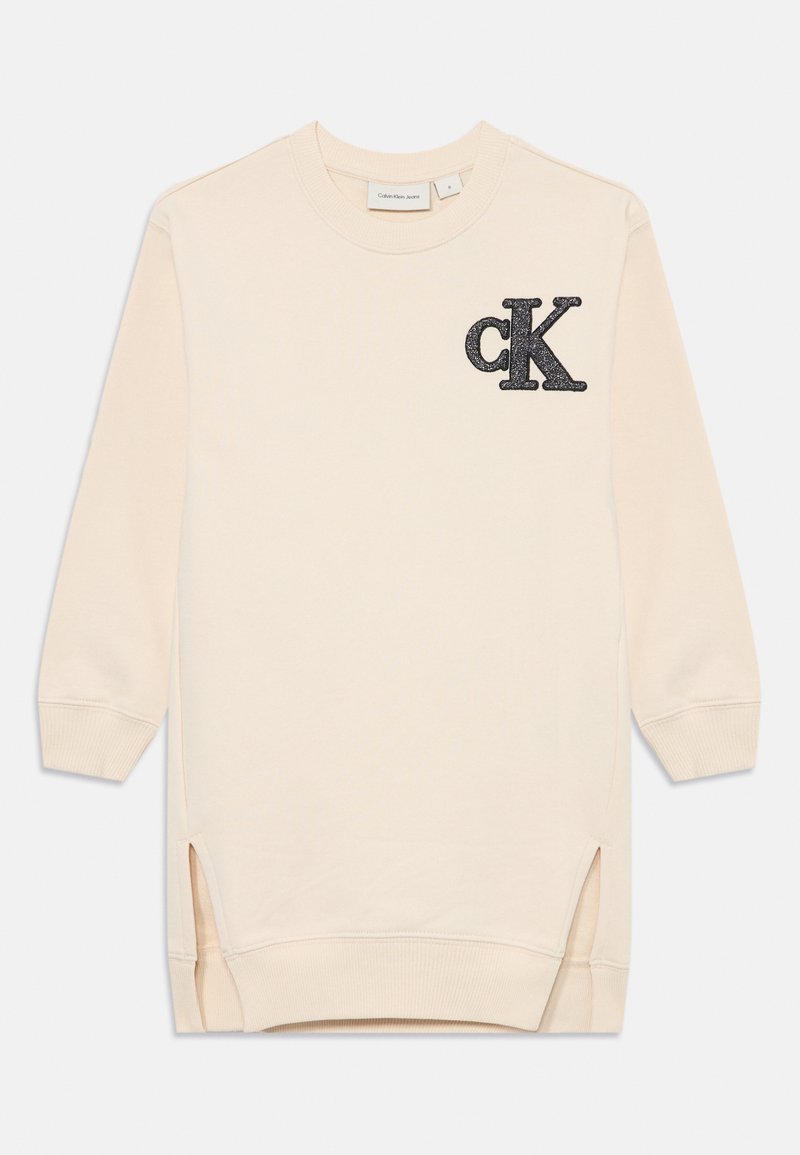 Lys beige genser med lange ermer, sidesplitt, og en teksturert svart "ck" logo på venstre bryst. Mykt bomullsmateriale.