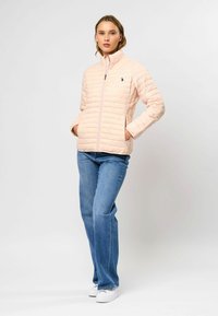 Vadderad, ljusrosa jacka med hög krage och dragkedja. Har en liten logotyp på bröstet, i kombination med blå utsvängda jeans och vita skor.