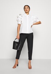 Camisa branca de botões com mangas bufantes, combinada com calças pretas risca de giz e uma mala preta tipo bucket. A modelo usa sandálias com salto transparente.