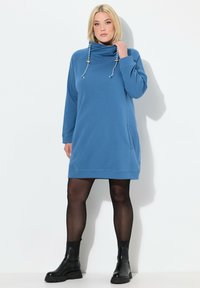 Blaues Sweatshirt-Kleid mit Kapuze, zwei seitlichen Taschen und dekorativen Kordeln. Es hat lange Ärmel und einen lässigen Schnitt. Kombiniert mit schwarzen Knöchelstiefeln.