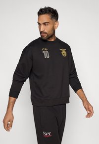 Felpa nera con dettagli ricamati in oro, inclusi il numero 10 e un logo. Presenta un collo e polsini a coste. Abbinata a pantaloni neri.