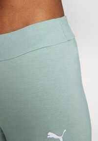 Tyrkysové legíny s hladkou, elastickou texturou a širokým pasem, s bílým logem Puma blízko dolní části nohy.