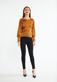 Een oranjetint met zwarte luipaardprint langemouwen top, gecombineerd met zwarte legging en puntige hakken, wat zorgt voor een getailleerd silhouet.