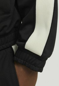 Sort sportsjakkeærme med en struktureret hvid stribe. Har elastiseret kant og glat stof. Minimalistisk design, nærbillede.