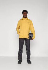 Κίτρινο αδιάβροχο μπουφάν με κουκούλα, συνδυασμένο με μαύρο παντελόνι και παπούτσια. Το άτομο κρατάει ένα μαύρο κράνος ποδηλάτου.