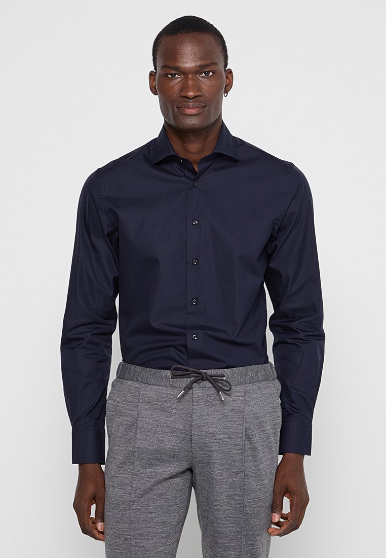 Granatowa koszula zapinana na guziki z długimi rękawami, usztywnionym kołnierzem i gładkiego materiału; zestawiona z szarymi spodniami dresowymi z troczkami i kieszeniami bocznymi.