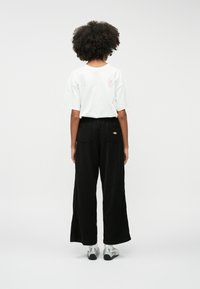 Egy göndör hajú személy áll háttal, fehér, oversized pólót, fekete, széles szárú nadrágot, fehér zoknit és ezüst színű sportcipőt visel.