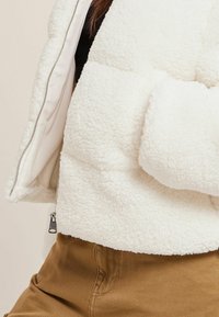 Chaqueta de forro polar blanca con superficie texturizada, cierre de cremallera y cuello alto. La tela es suave, con un diseño acolchado que enfatiza la calidez.