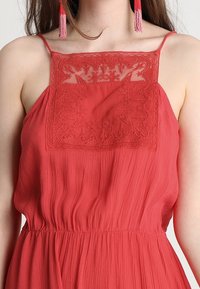 Robe rouge avec un corsage texturé et brodé comportant des motifs floraux complexes, des bretelles fines et une jupe fluide et froncée.