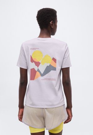 Άτομο με κοντά μαλλιά που φορά ανοιχτόχρωμο T-shirt με πολύχρωμο γραφικό βουνό και κείμενο στην πλάτη, συνδυασμένο με μπεζ σορτς και κίτρινες κολάν.