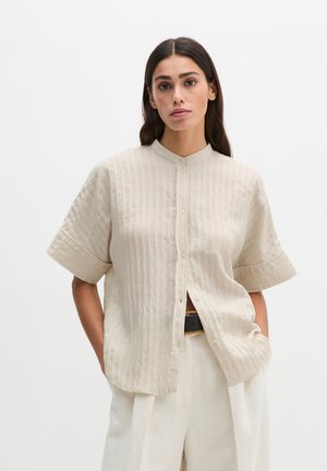 Femme aux longs cheveux noirs portant une chemise boutonnée texturée à manches courtes beige et un pantalon blanc ample, les mains dans les poches.