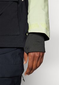 Maniche di giacca nere e verde chiaro con evidenti dettagli in velcro. Sottogiacca nera e aderente. Texture liscia con blocchi di colore a contrasto.