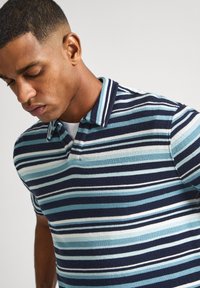 Svītrota polo krekls ar maiņojošām tumši zila, gaiši zila un baltām svītrām. Mīksta tekstūra ar klasisku nolokāmu apkakli un īsām piedurknēm.