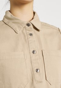 Gros plan d'une chemise beige à boutons avec col et deux poches poitrine, présentant quatre boutons-pression argentés.