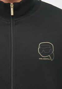 Μαύρο μπουφάν με φερμουάρ και ψηλό γιακά. Διαθέτει χρυσ�ό φερμουάρ και χρυσό λογότυπο με στυλιζαρισμένο πρόσωπο που φορά γυαλιά ηλίου στο αριστερό στήθος.