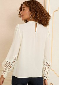 White blouse with long sleeves featuring intricate lace trim at the cuffs and a small keyhole detail at the back. Smooth, lightweight fabric.