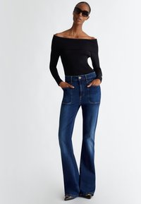Top nero off-the-shoulder con maniche lunghe, abbinato a jeans a zampa d'elefante blu scuro con grandi tasche anteriori e cuciture a contrasto.