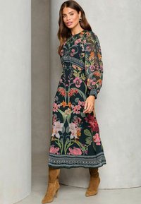 Floral midi mekko, jossa on tumma tausta värikkäine kukkineen, läpikuultavat pitkät hihat ja brodeeratut yksityiskohdat vyötäröllä ja helman kohdalla.