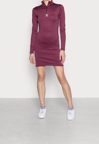 Abito lungo a maniche lunghe color marsala con colletto con zip e design aderente; presenta un logo bianco sul petto. Abbinato a sneakers bianche.