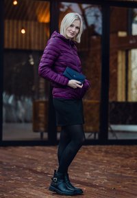 Purple puffer jacket, black skirt, black tights, and ankle boots with buckle. Holding a blue clutch. Wooden floor and glass backdrop.