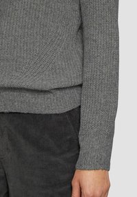 Grau gestrickter Pullover mit einer strukturierten, dicken Webart, mit einem gerippten Saum und Bündchen. Getragen über dunklen Cordhosen.