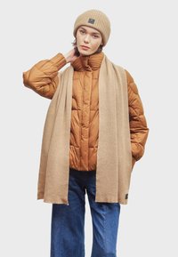 Bruine pufferjack met een hoge kraag, beige geribbelde beanie en een lange beige sjaal. Donkerdenim wijde broek. Zwarte logoaccenten.