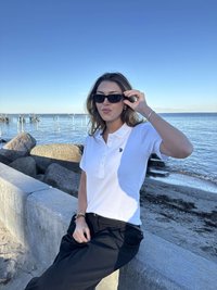 Biała, prążkowana koszulka polo z plisą na guziki i logo. Czarne, luźne spodnie. Czarne okulary przeciwsłoneczne. Nadmorski krajobraz z skałami i wodą.