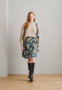 Beige ribbestrikket genser, teal blomstrete satengskjørt, svarte knelang støvler og en liten svart veske. Modellen står mot en nøytral bakgrunn.