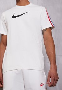 Bílý bavlněný tričko s černým logem Nike na prsou a červenobílými pruhovanými detaily na rukávech, s volným střihem.