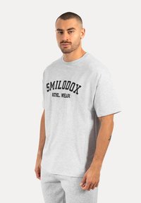 Camiseta gris de manga corta hecha de algodón, con letras en negrita negras que dicen "SMILODOX ATHL. WEAR" en la parte frontal. Diseño clásico de cuello redondo.