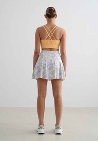 Sport-BH in hellorange mit überkreuzten Trägern, kombiniert mit einem ausgestellten Rock im pastellfarbenen Blumendesign. Das Model trägt weiße Sneaker.