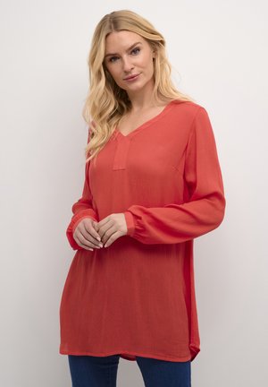 Tunique à col en V, à manches longues, de couleur corail éclatant, fabriquée en tissu texturé avec une coupe décontractée et des fentes sur les côtés, associée à un jean foncé.