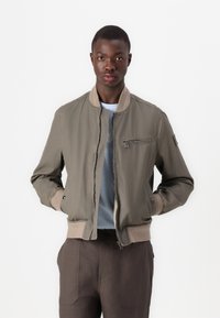 Jachetă bomber khaki cu un buzunar cu fermoar, manșete și tiv cu elastic. Materialul are o textură netedă; purtată peste o cămașă albastră deschisă și pantaloni închiși la culoare.