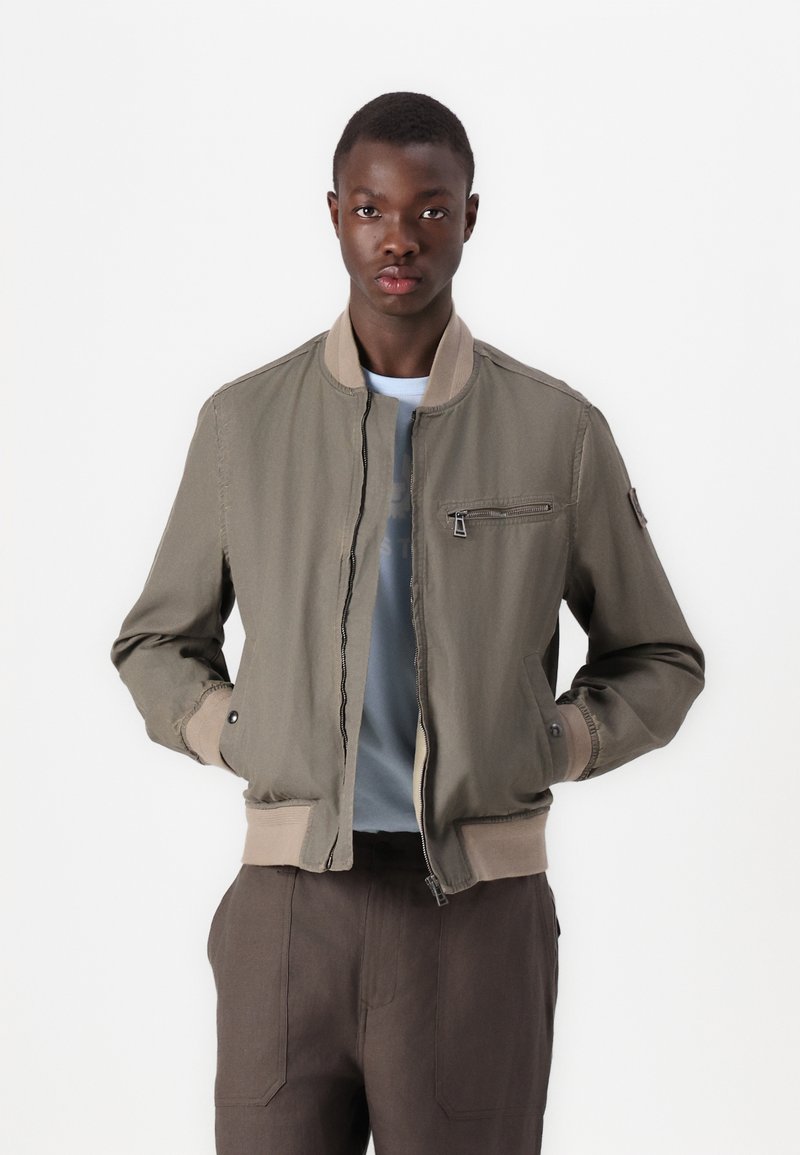 Giacca bomber khaki con tasca con cerniera, polsini e orlo a coste. Il materiale ha una texture liscia; indossata sopra una camicia azzurra chiara e pantaloni scuri.