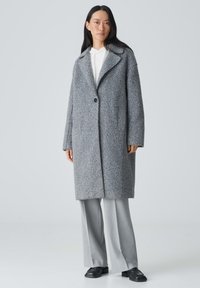 Grauer Wollmantel mit eingelegtem Kragen, einzelnen Knopfverschluss und seitlichen Taschen, kombiniert mit grauen Hosen und schwarzen Schuhen.