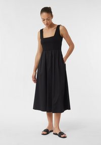 Schwarzes ärmelloses Kleid mit quadratischem Ausschnitt, tailliertem Design, fließendem Rock und seitlichen Taschen; glatter Stoff und knöchellange Ausführung.