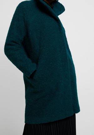 Person trägt einen türkisfarbenen, strukturierten Wollmantel mit hohem Kragen und Seitentaschen, kombiniert mit einem schwarzen plissierten Rock vor einfachem Hintergrund.